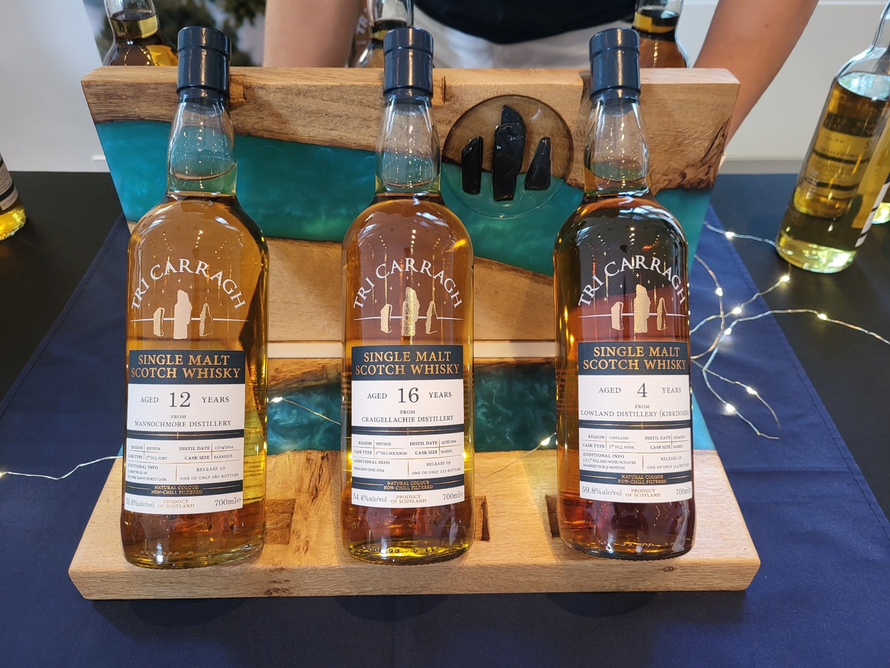 The Trio — bespoke oak and Belhaven Teal resin whisky bottle display holding three Tri Carragh single malt bottles at 45 degrees, LED-lit backboard with slate three-pillar logo